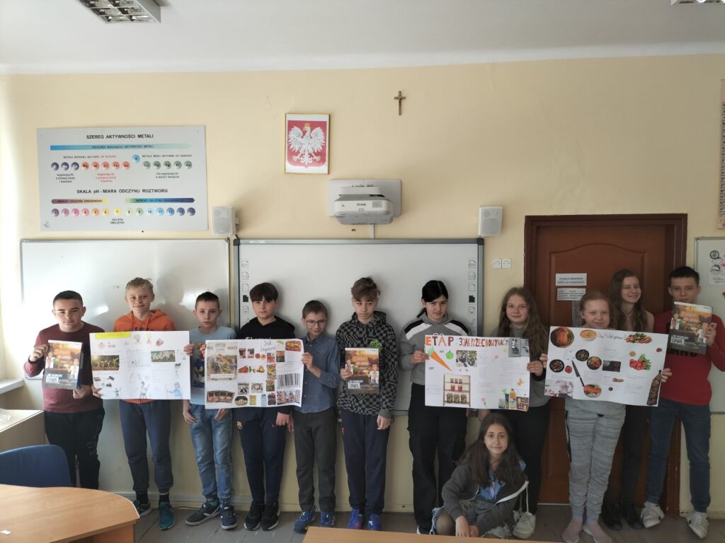Zdjęcie przedstawia uczniów stojących pod tablicą prezentujących plakaty.