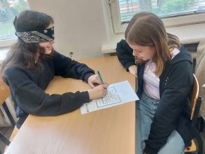 Zdjęcie przedstawia jak uczennica rozwiązuje zadanie z zamkniętymi oczyma kierowana przez koleżankę