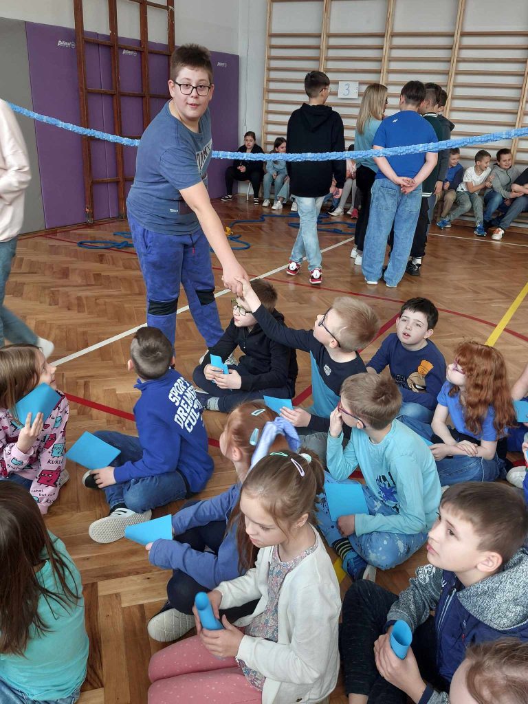 zdjęcie przedstawia jak uczniowie siedzą na podłodze w sali gimnastycznej