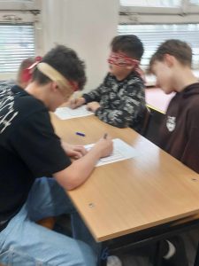 zdjęcie przedstawia uczeń rozwiązuje zadania z zamkniętymi oczyma kierowany przez kolegę.