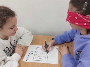 Zdjęcie przedstawia jak uczennica rozwiązuje zadanie z zamkniętymi oczyma kierowana przez koleżankę