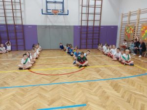 dzieci siedzą na podłodze w rządku podzieleni na grupy.