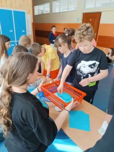 Zdjęcie przedstawia ucznia losującego niebieską kopertę z hasłem z czerwonego koszyczka