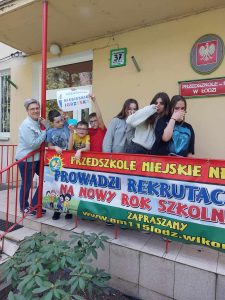 Zdjęcie przedstawia uczniów stojących przed przedszkolem z plakatem niebieskie igrzyska