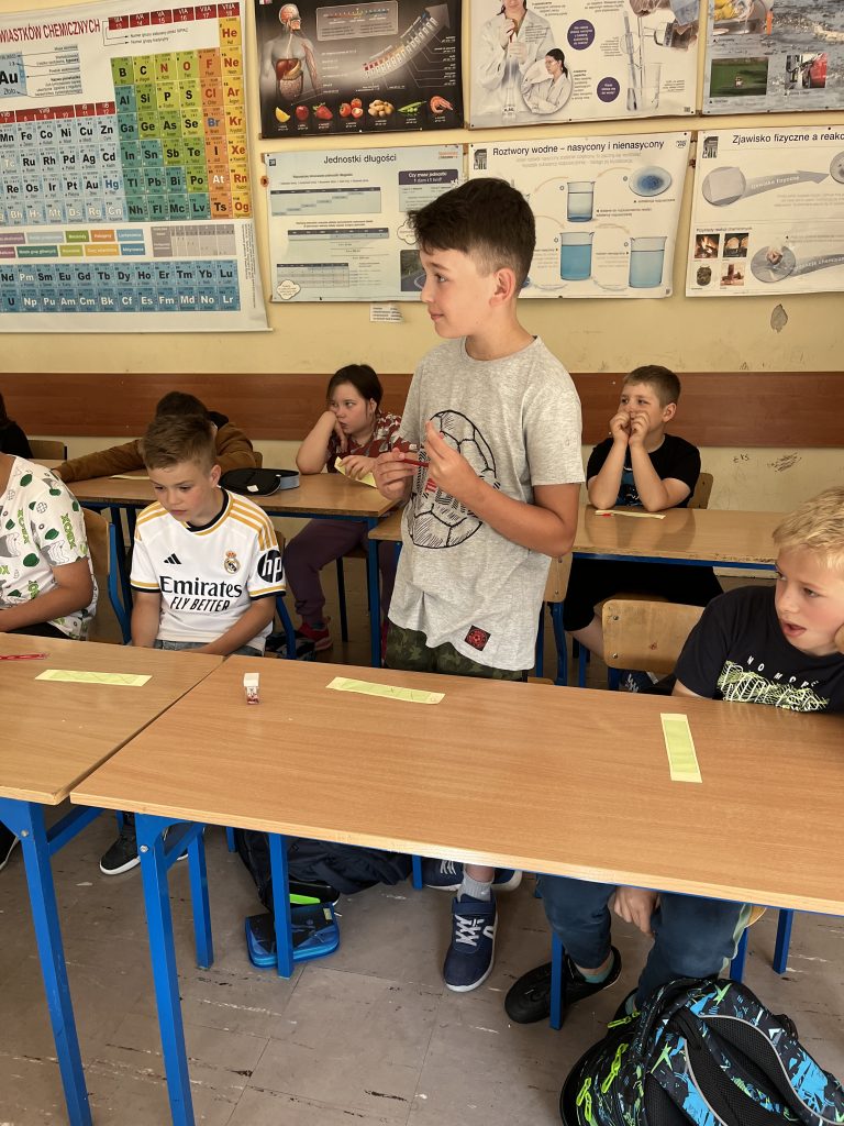 uczniowie stoją przy ławeczkach, przed nimi leżą karteczki z namalowanymi szansami, na głównym planie zwycięzca