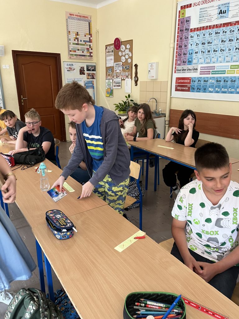 uczniowie stoją przy ławeczkach, przed nimi leżą karteczki z namalowanymi szansami