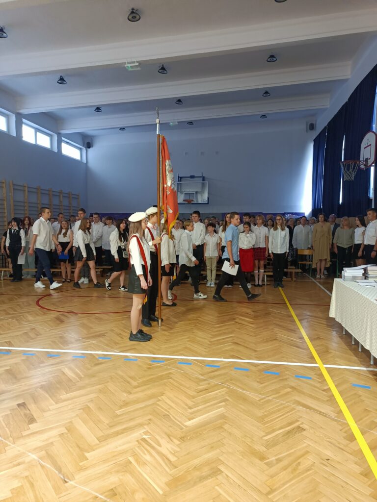zdjęcie uczniów w sztandarze na tle uczniów klas ósmych i siódmych 
