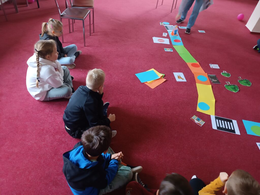 Dzieci siedzą na dywanie i zgłaszają się do odpowiedzi, przed nimi znajduje się wieloformatowa gra planszowa.