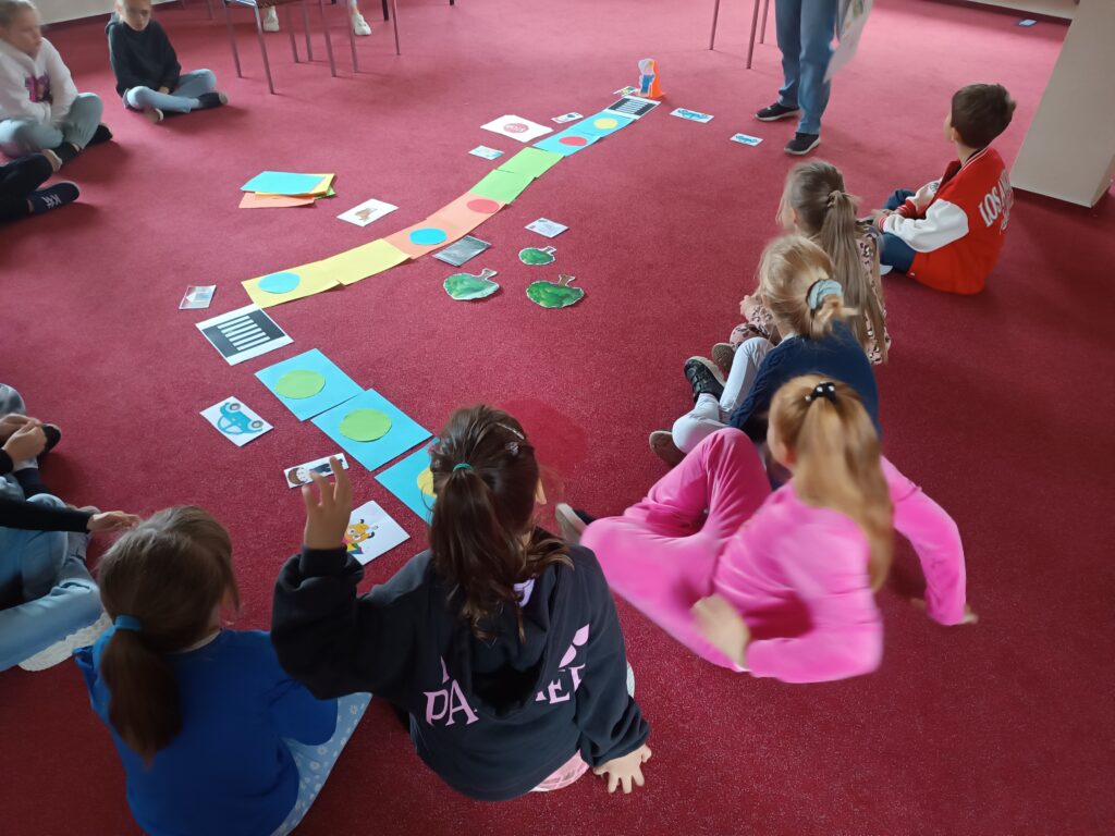 Dzieci siedzą na dywanie i zgłaszają się do odpowiedzi, przed nimi znajduje się wieloformatowa gra planszowa.