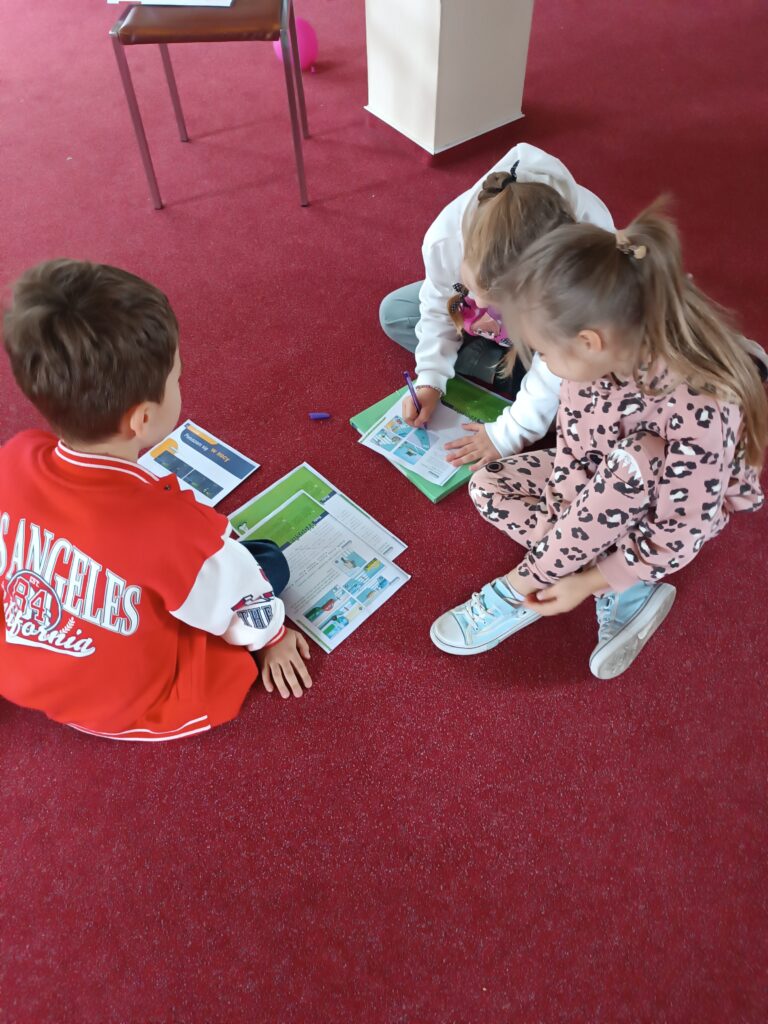 Dzieci siedzą na dywanie i zgłaszają się do odpowiedzi, przed nimi znajduje się wieloformatowa gra planszowa.