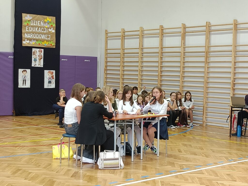 Na pierwszym planie uczennicę siedzące przy stole odgrywają rolę nauczycielek w pokoju nauczycielskim