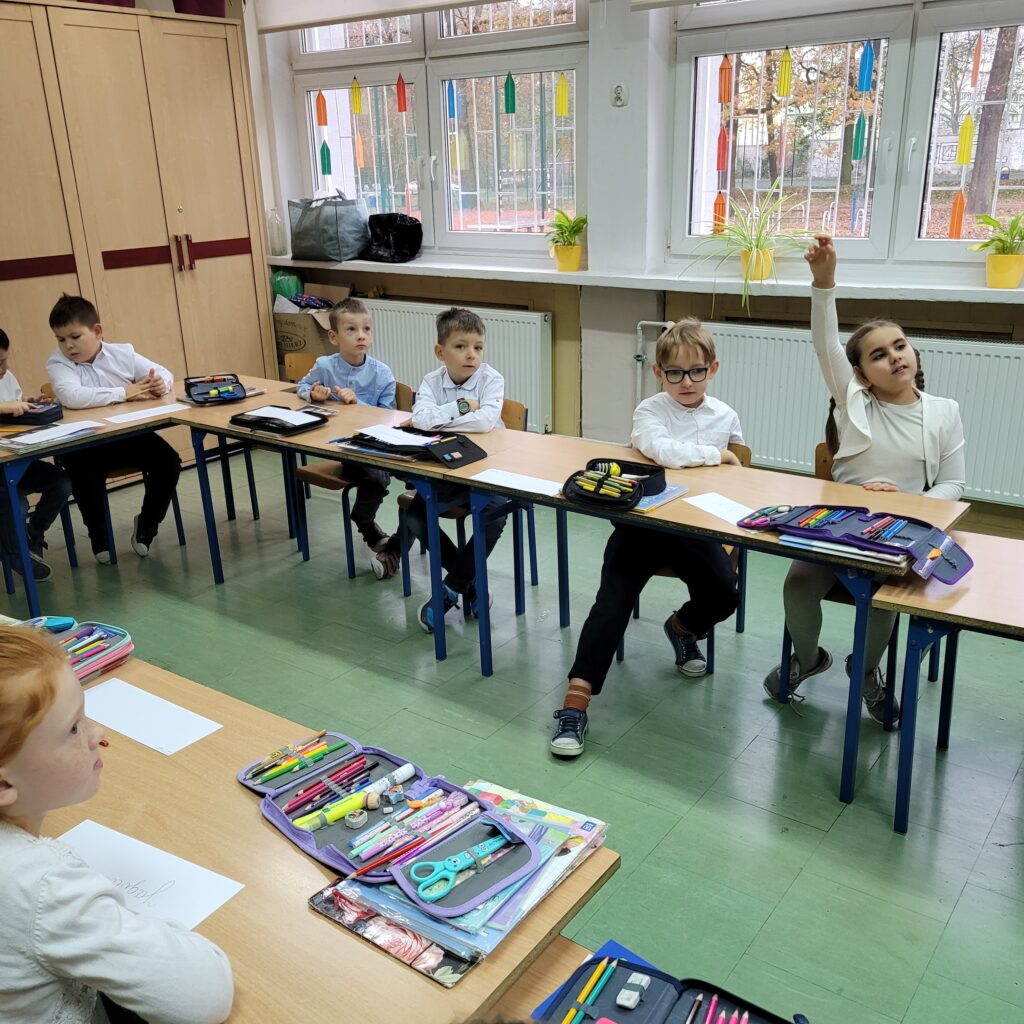 dzieci słuchają, jedna z dziewczynek trzyma rękę do góry chcąc się zgłosić