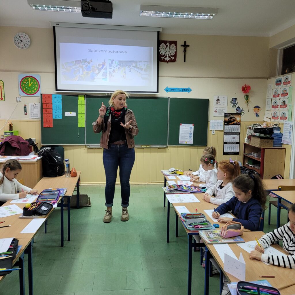 pani na środku sali tłumaczy coś dzieciom siedzącym w ławkach