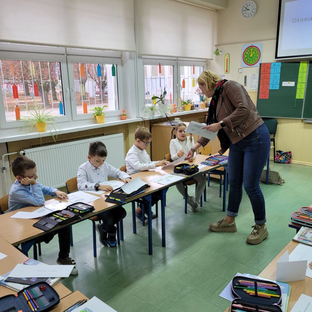 pani rozdaje uczniom siedzącym w ławkach karty pracy