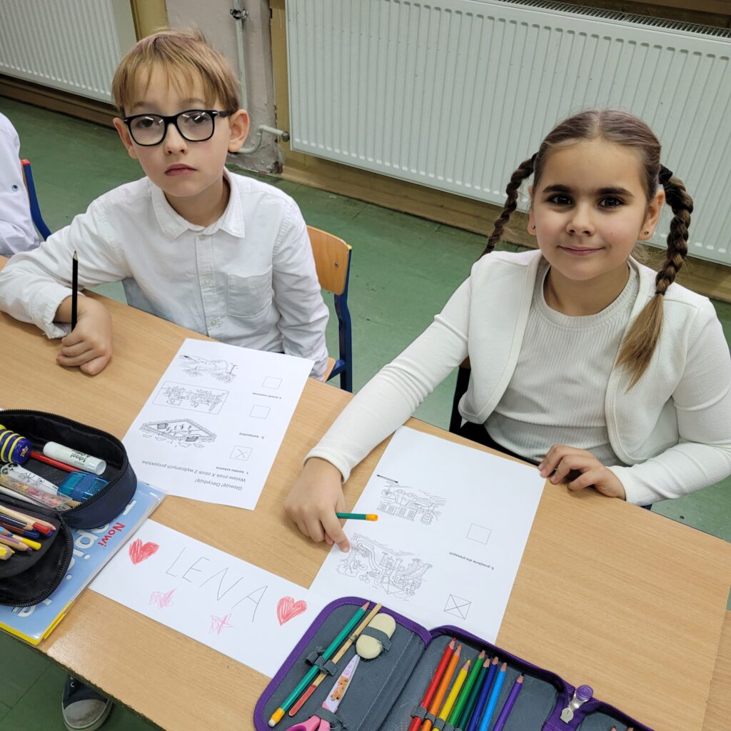 uczniowie siedzą w ławce przy kartach pracy, patrzą w obiektyw