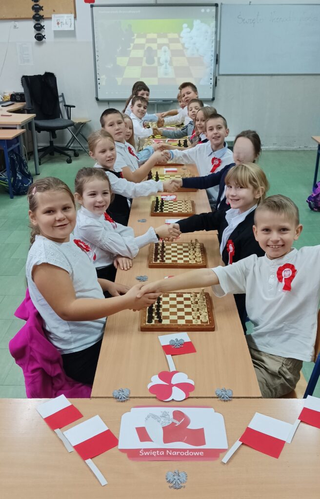 uczniowie siedzą przy szachach wokół flagi białoczerwone i dzieci mają przypięte kotyliony