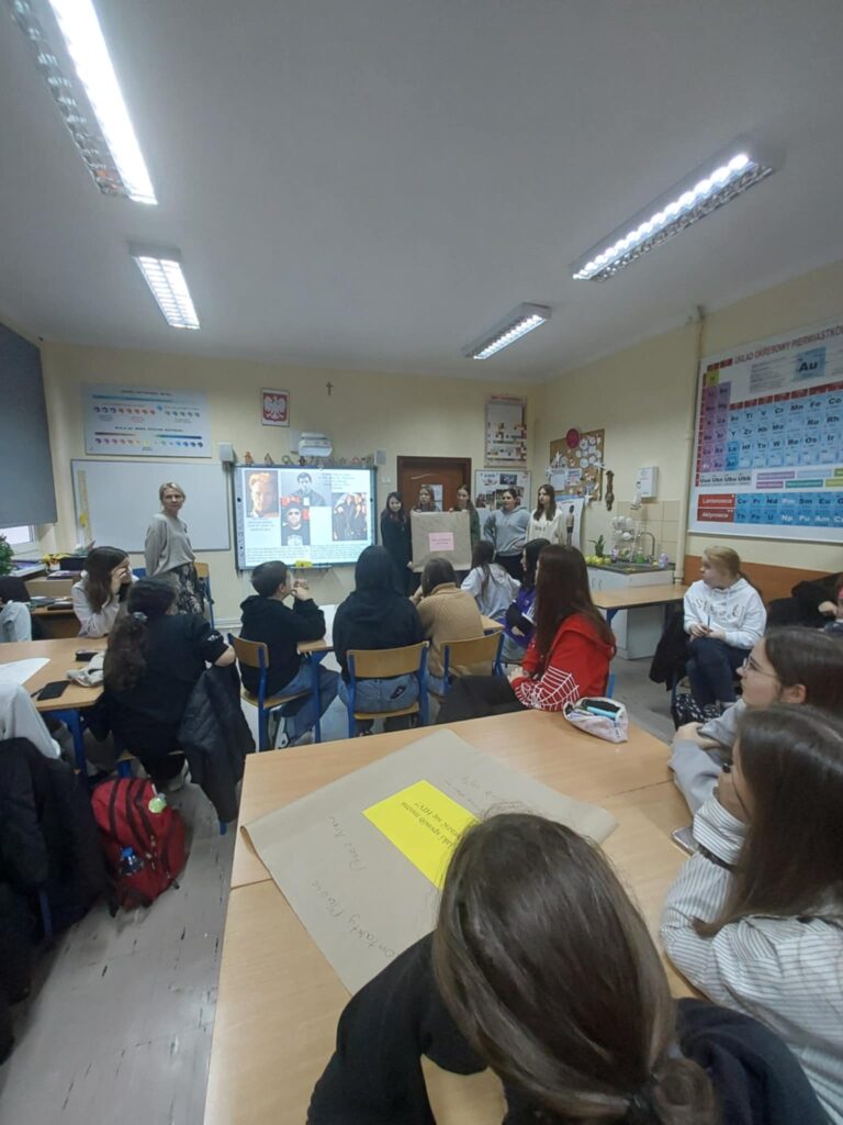 Na zdjęciu widać uczniów i uczennice siedzące przy stolikach, a w tle grupę referującą wykonane zadanie i słuchającą panią pedagog.