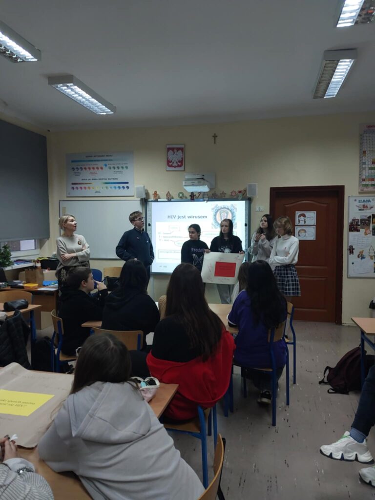 Na zdjęciu widać uczniów i uczennice siedzące przy stolikach, a w tle grupę referującą wykonane zadanie.