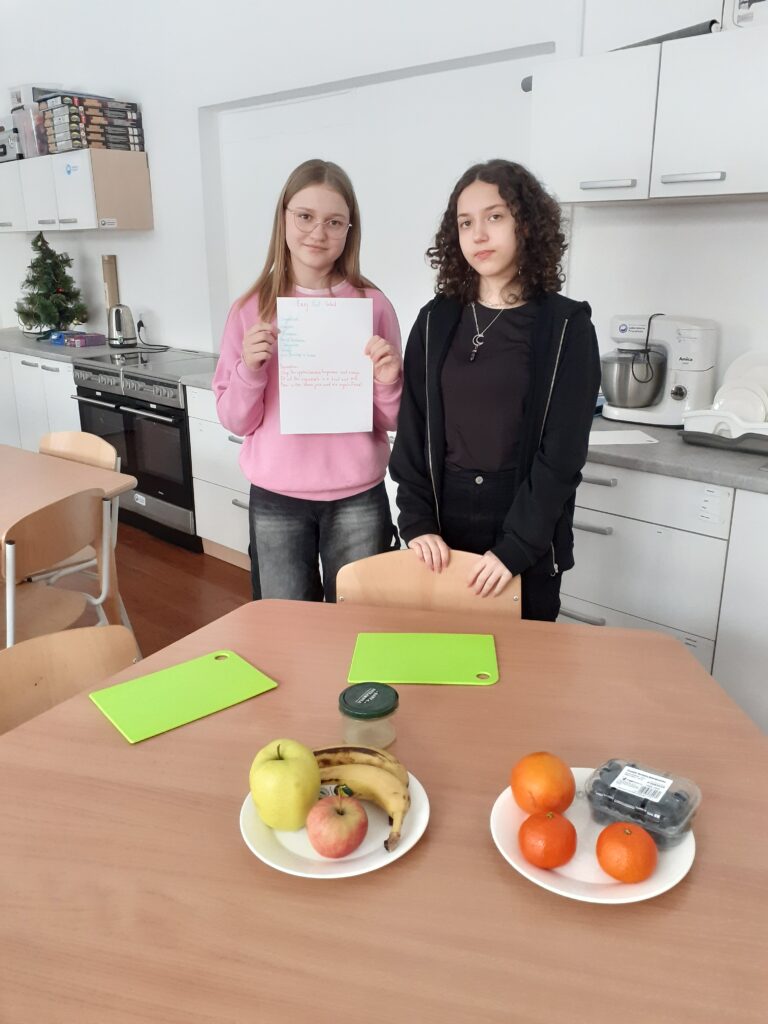 na zdjęciu widzimy dwie uczennice stojące przy stole, na którym leżą owoce. Jedna z uczennic trzyma plakat, prezentując przepis na sałatkę owocową.
