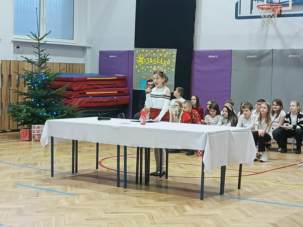 uczennica przemawia przy stole z białym obrusem na scenie