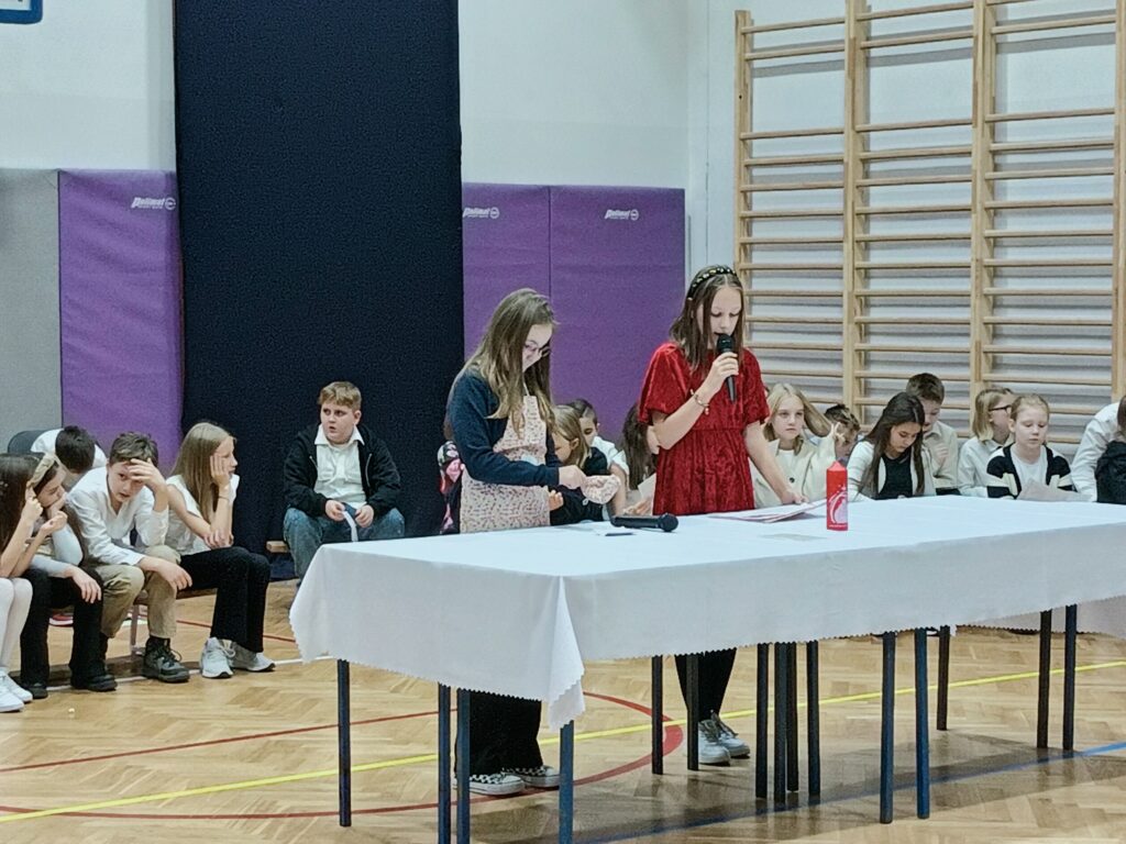 dwie uczennice stoją przy stole z białym obrusem na scenie
