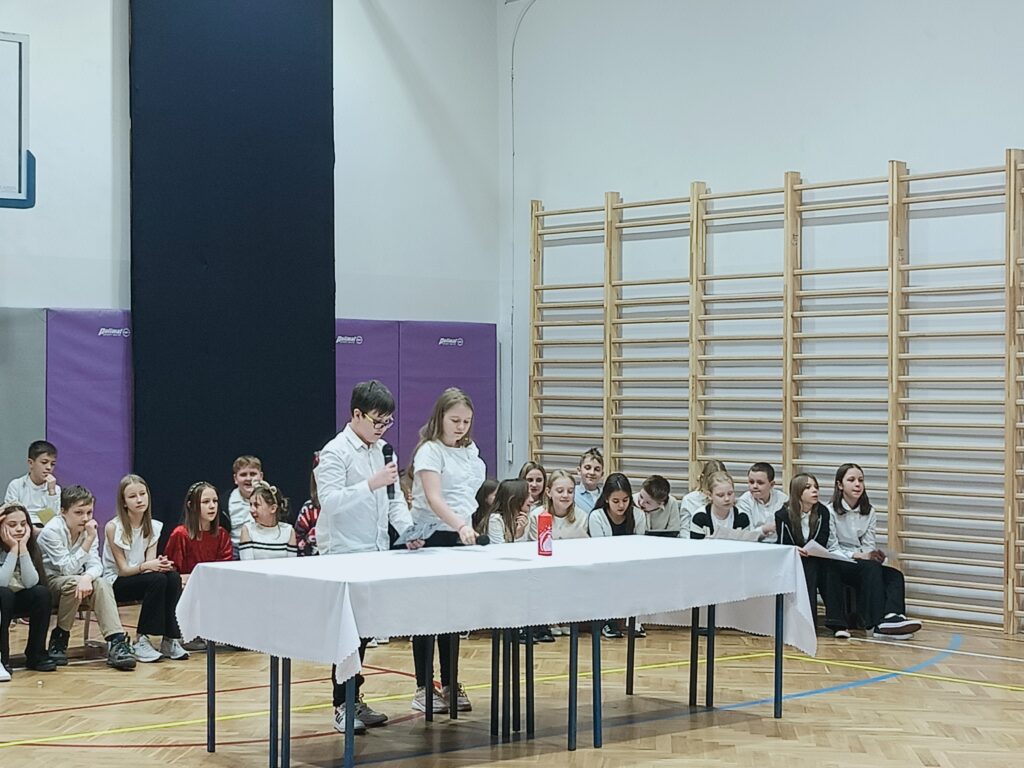 dwoje uczniów stoi przy stole z białym obrusem na scenie