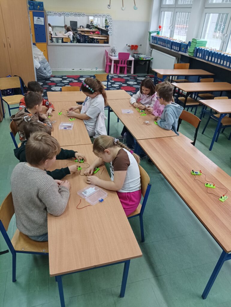 dzieci siedzą przy ławkach i wykonują doświadczenia z prądem