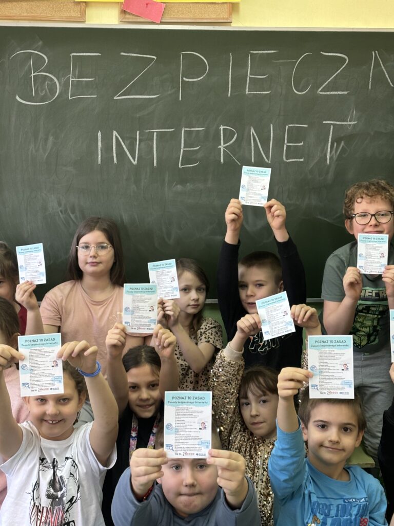 uczniowie siedzą przed tablicą trzymają w rączkach ulotki z DBI w tle napis na tablicy Bezpieczny Internet