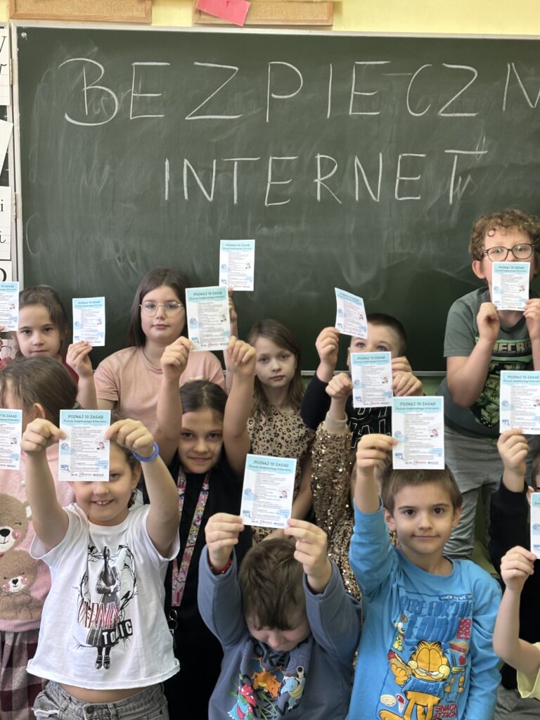 uczniowie siedzą przed tablicą trzymają w rączkach ulotki z DBI w tle napis na tablicy Bezpieczny Internet