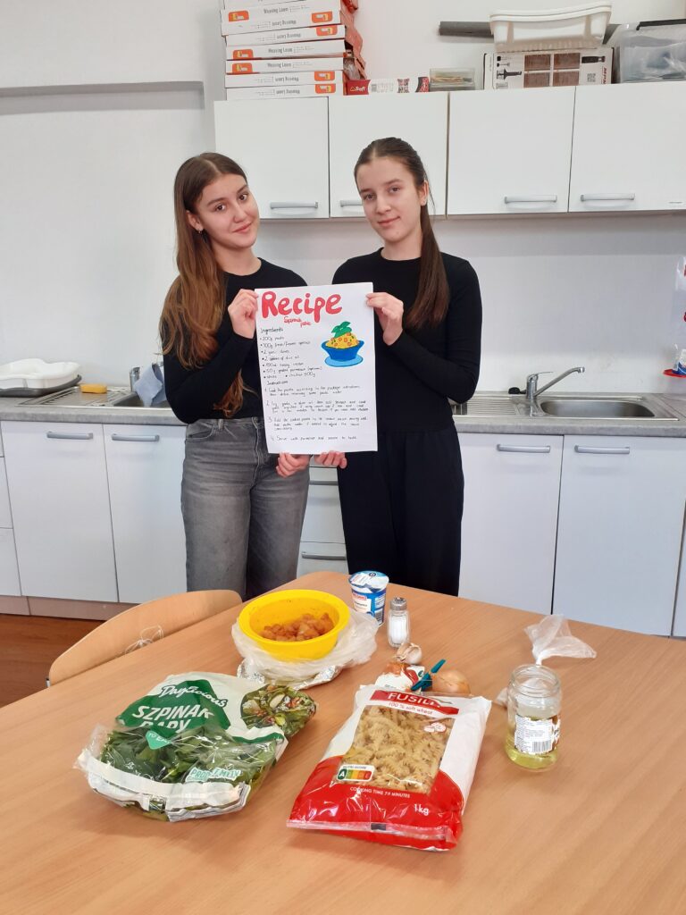 na zdjęciu widzimy dwie uczennice trzymające plakat z przepisem na potrawę. Stoją one przy stole, na którym wyłożone są produkty spożywcze używane w przepisie.