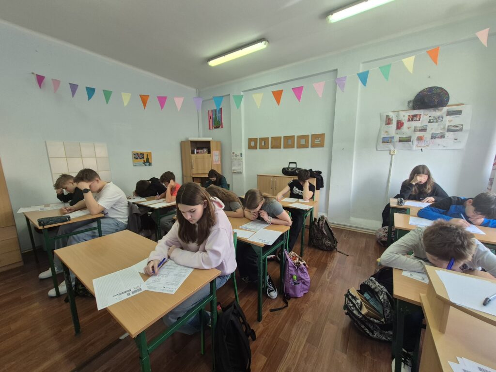 uczniowie siedzą przy ławkach z kartami zadań i rozwiązują zadania konkursowe