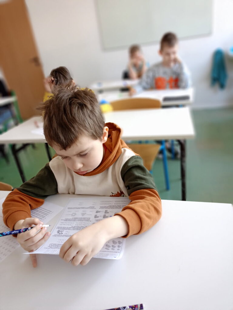 uczniowie siedzą przy ławkach z kartami zadań i rozwiązują zadania konkursowe