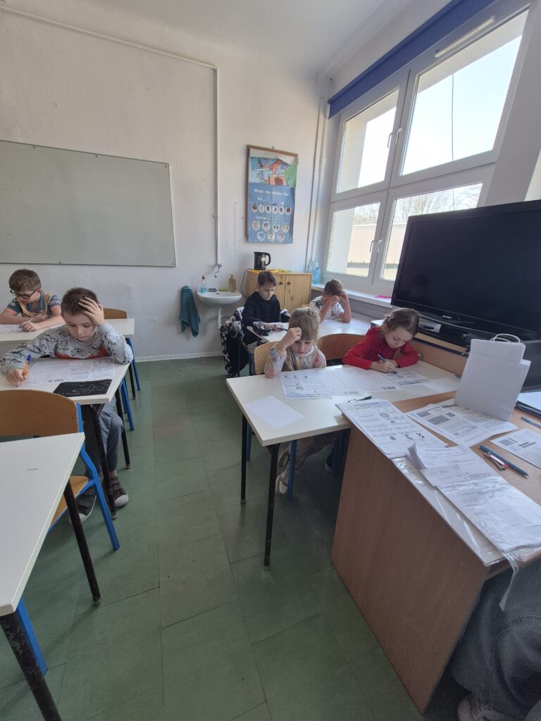 uczniowie siedzą przy ławkach z kartami zadań i rozwiązują zadania konkursowe