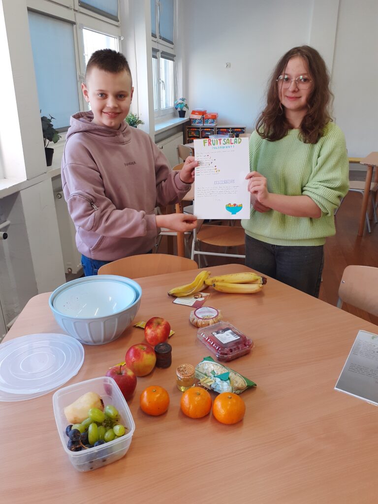 na zdjęciu widzimy ucznia i uczennicę trzymających plakat z przepisem na sałatkę owocową. Stoją oni przy stole, na którym wyłożone są produkty spożywcze używane w przepisie.