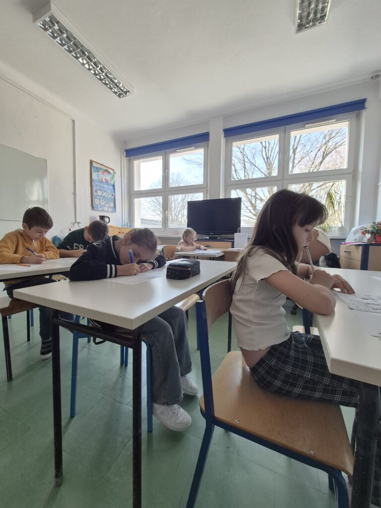 uczniowie siedzą przy ławkach z kartami zadań i rozwiązują zadania konkursowe