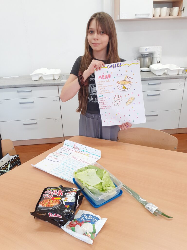 na zdjęciu widzimy uczennicę trzymającą plakat z przepisem na potrawę, stojącą przy stole, na którym wyłożone są produkty spożywcze używane w przepisie.