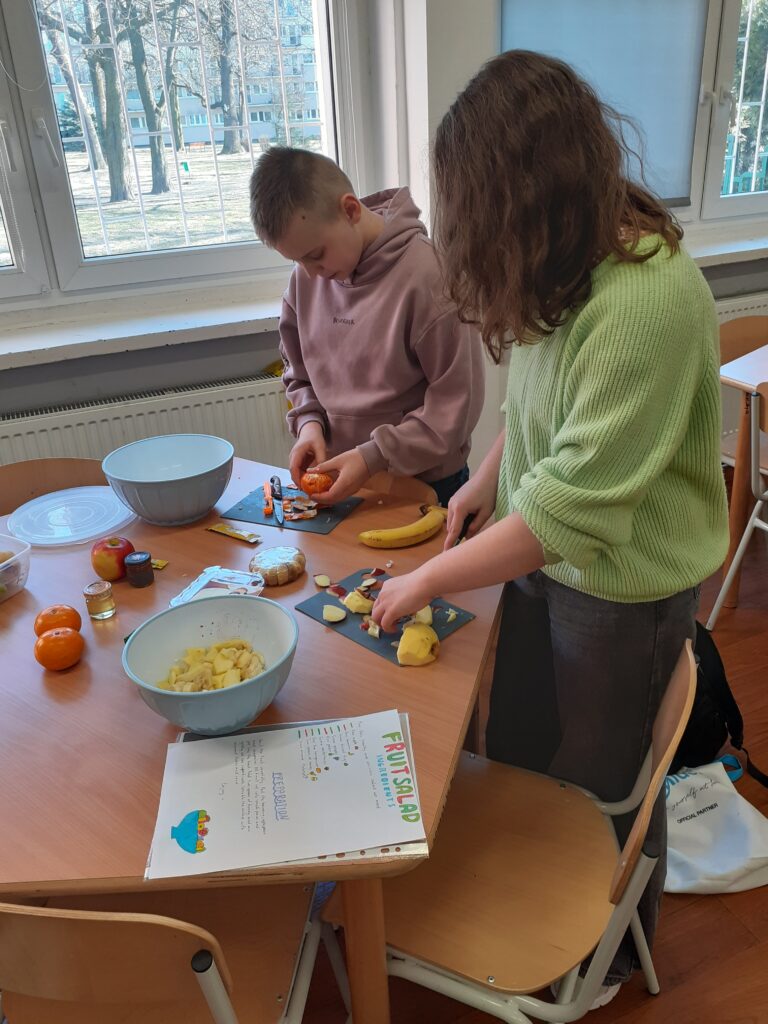na zdjęciu widzimy ucznia i uczennicę stojących przy stoliku i obierających lub krojących składniki na sałatkę owocową.