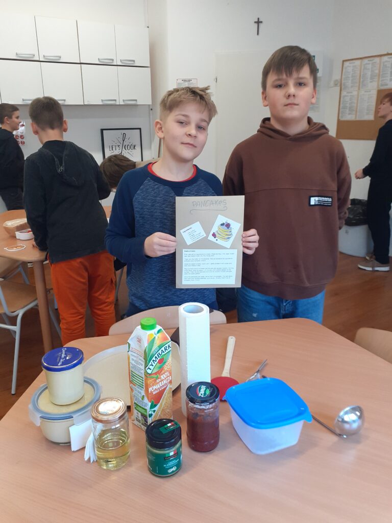 na zdjęciu widzimy dwóch uczniów trzymających plakaty z przepisami na potrawy. Stoją oni przy stole, na którym wyłożone są produkty spożywcze używane w przepisie.