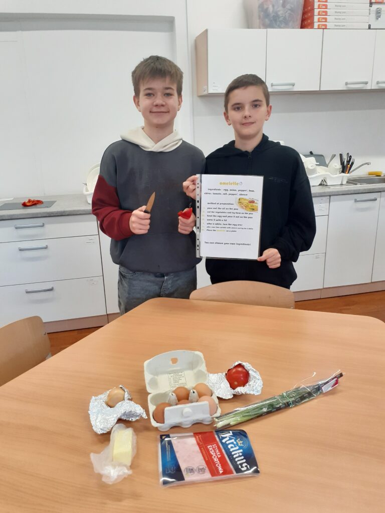 na zdjęciu widzimy dwóch uczniów trzymających plakaty z przepisami na potrawy. Stoją oni przy stole, na którym wyłożone są produkty spożywcze używane w przepisie.