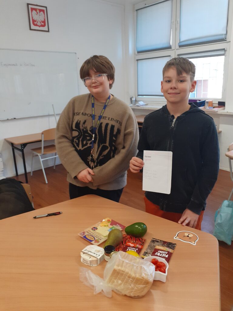 na zdjęciu widzimy dwóch uczniów trzymających plakaty z przepisami na potrawy. Stoją oni przy stole, na którym wyłożone są produkty spożywcze używane w przepisie.