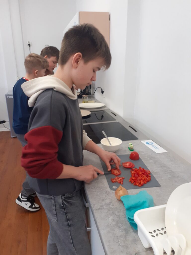 na zdjęciu widzimy uczniów stojących przy blacie kuchennym i kuchence, przygotowujących swoje dania.