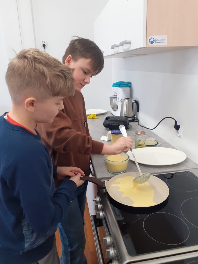 na zdjęciu widzimy uczniów stojących przy blacie kuchennym i kuchence, przygotowujących swoje dania.