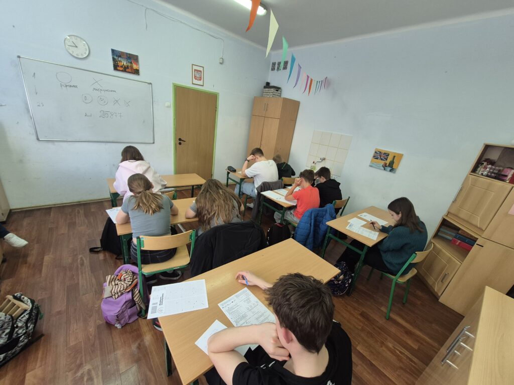 uczniowie siedzą przy ławkach z kartami zadań i rozwiązują zadania konkursowe