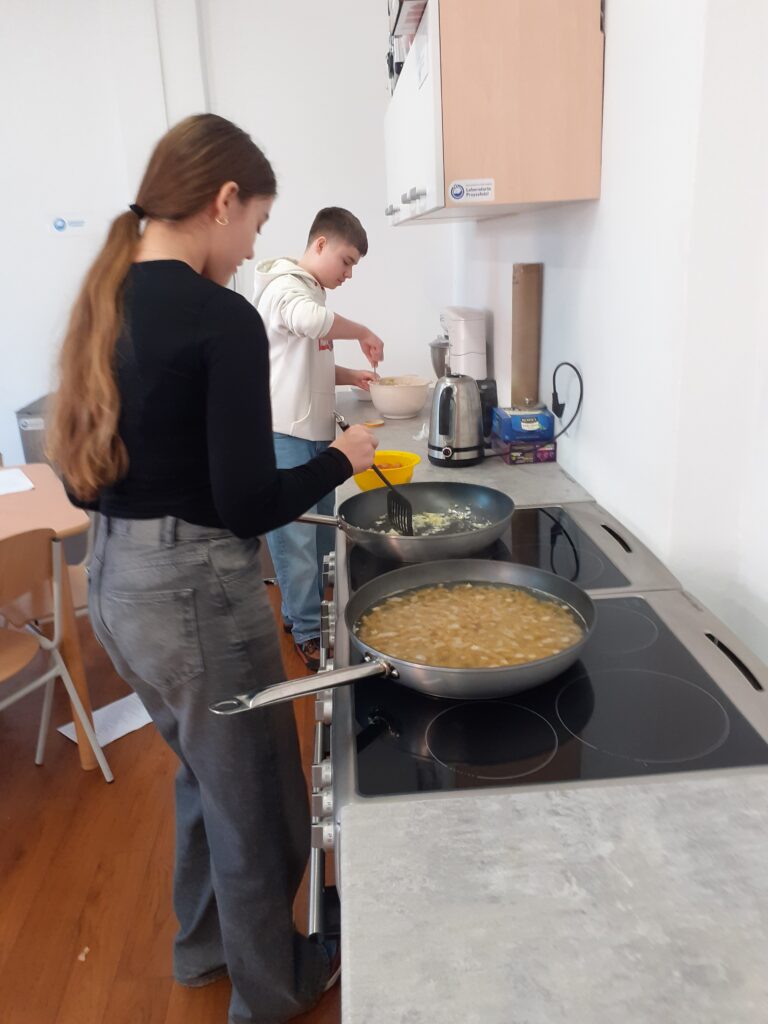 na zdjęciu widzimy uczniów stojących przy blacie kuchennym i kuchence, przygotowujących swoje dania.