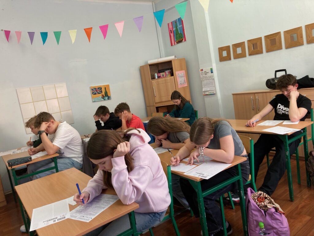 uczniowie siedzą przy ławkach z kartami zadań i rozwiązują zadania konkursowe