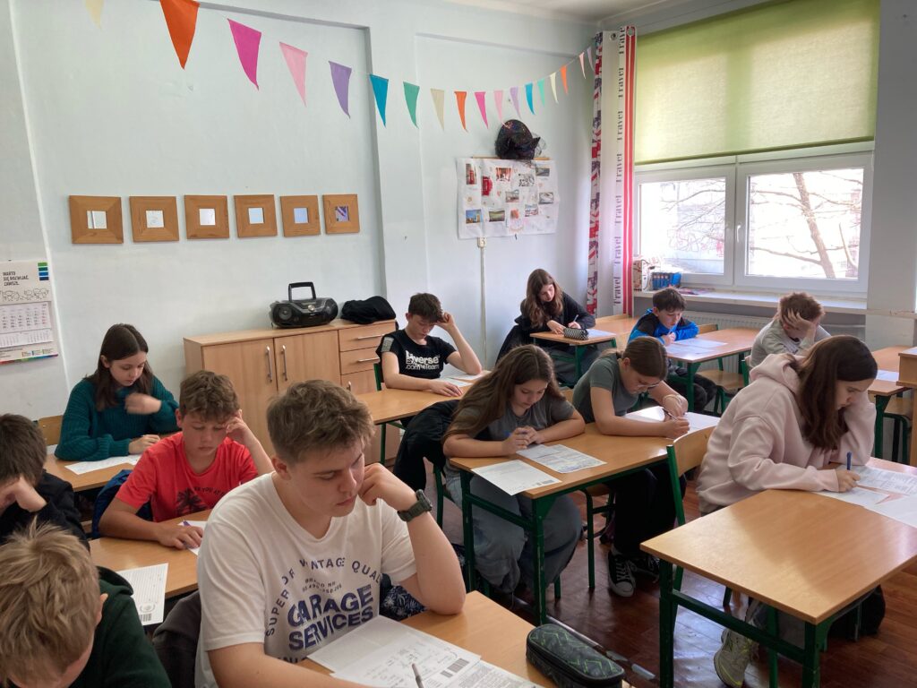 uczniowie siedzą przy ławkach z kartami zadań i rozwiązują zadania konkursowe