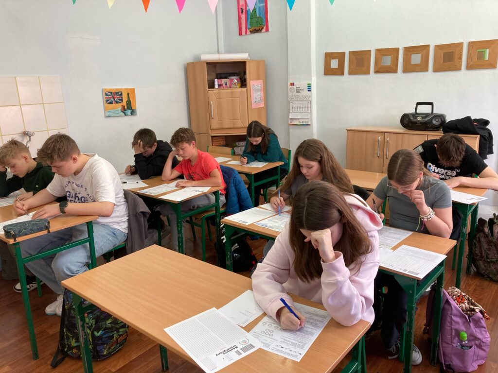 uczniowie siedzą przy ławkach z kartami zadań i rozwiązują zadania konkursowe