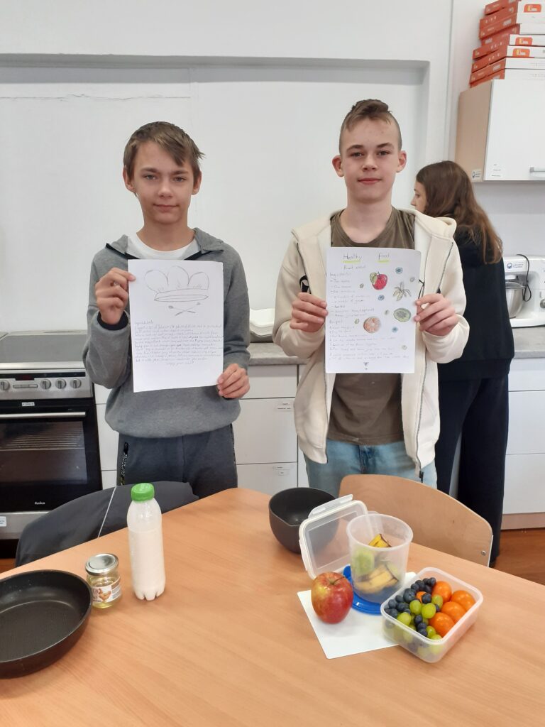na zdjęciu widzimy dwóch uczniów trzymających plakaty z przepisami na potrawy. Stoją oni przy stole, na którym wyłożone są produkty spożywcze używane w przepisie.
