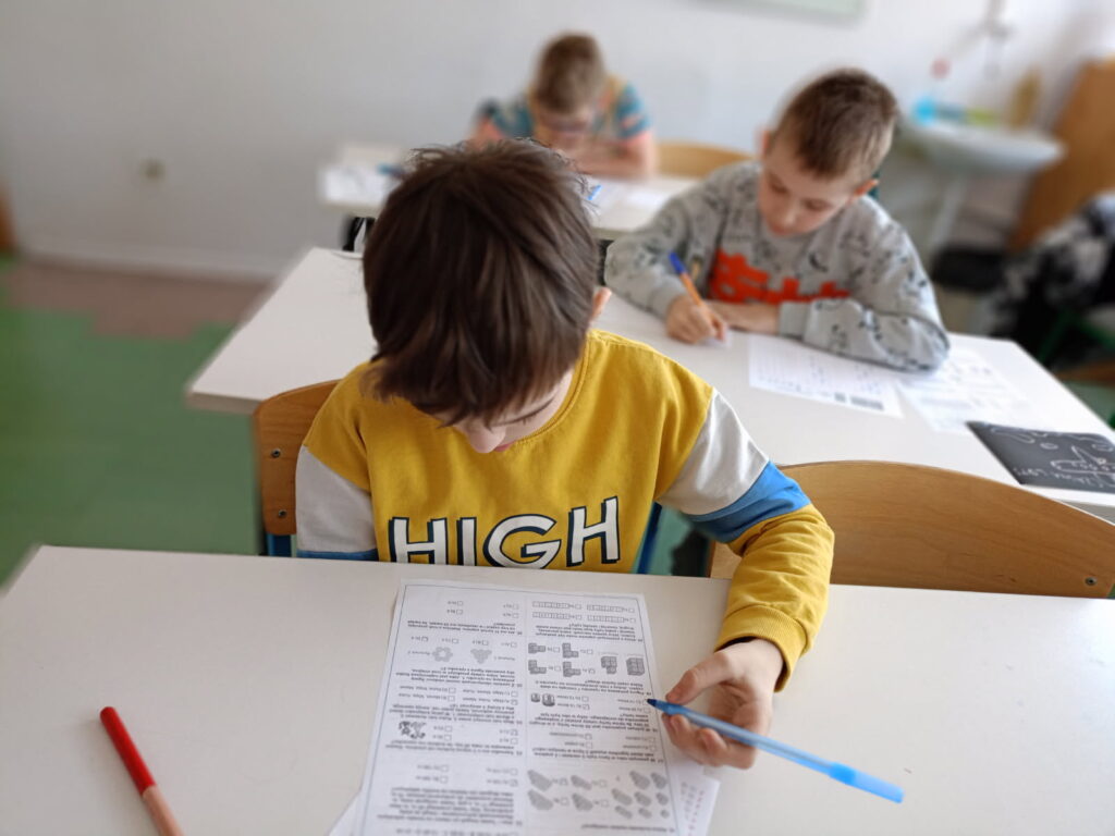 uczniowie siedzą przy ławkach z kartami zadań i rozwiązują zadania konkursowe