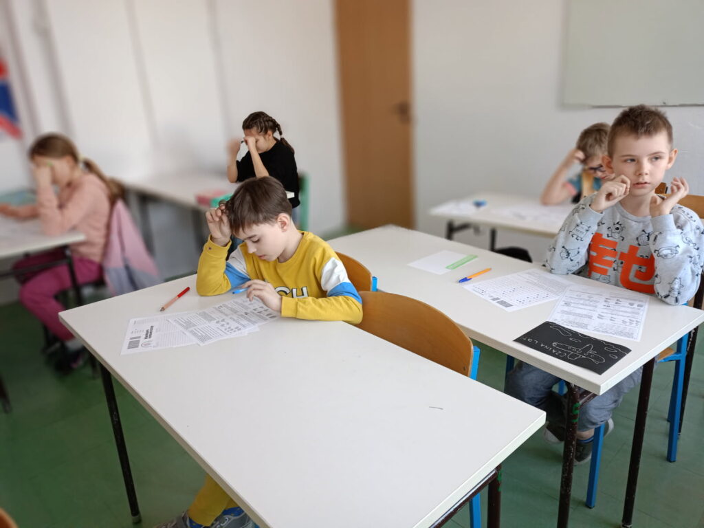 uczniowie siedzą przy ławkach z kartami zadań i rozwiązują zadania konkursowe